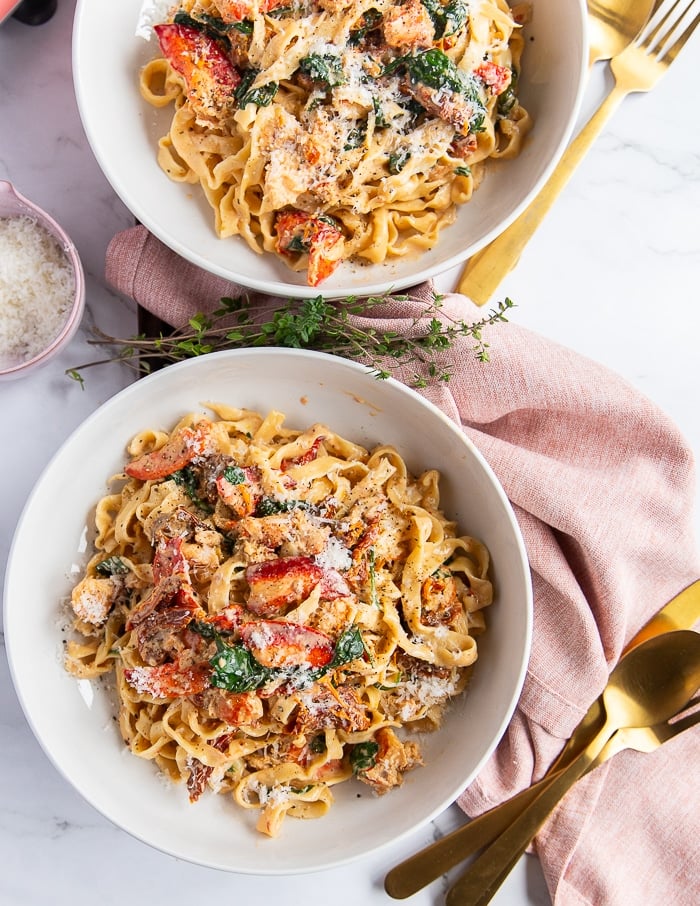 two lobster pasta bowls garnished with parmesan cheese and surrounded by golden cutlery. The pasta is creamy and divine with cooked lobster meat on top