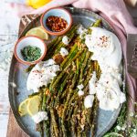 A plate of roasted asparagus with parmesan cheese served with some burrata cheese on the side, some extra chilli flakes for sprinkling, some herbs and parmesan cheese on top and lemon slices on the side