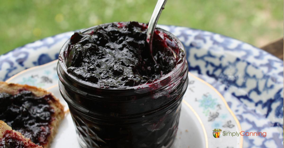 A full open canning jar of deeply colored blueberry jam sitting on a china plate.