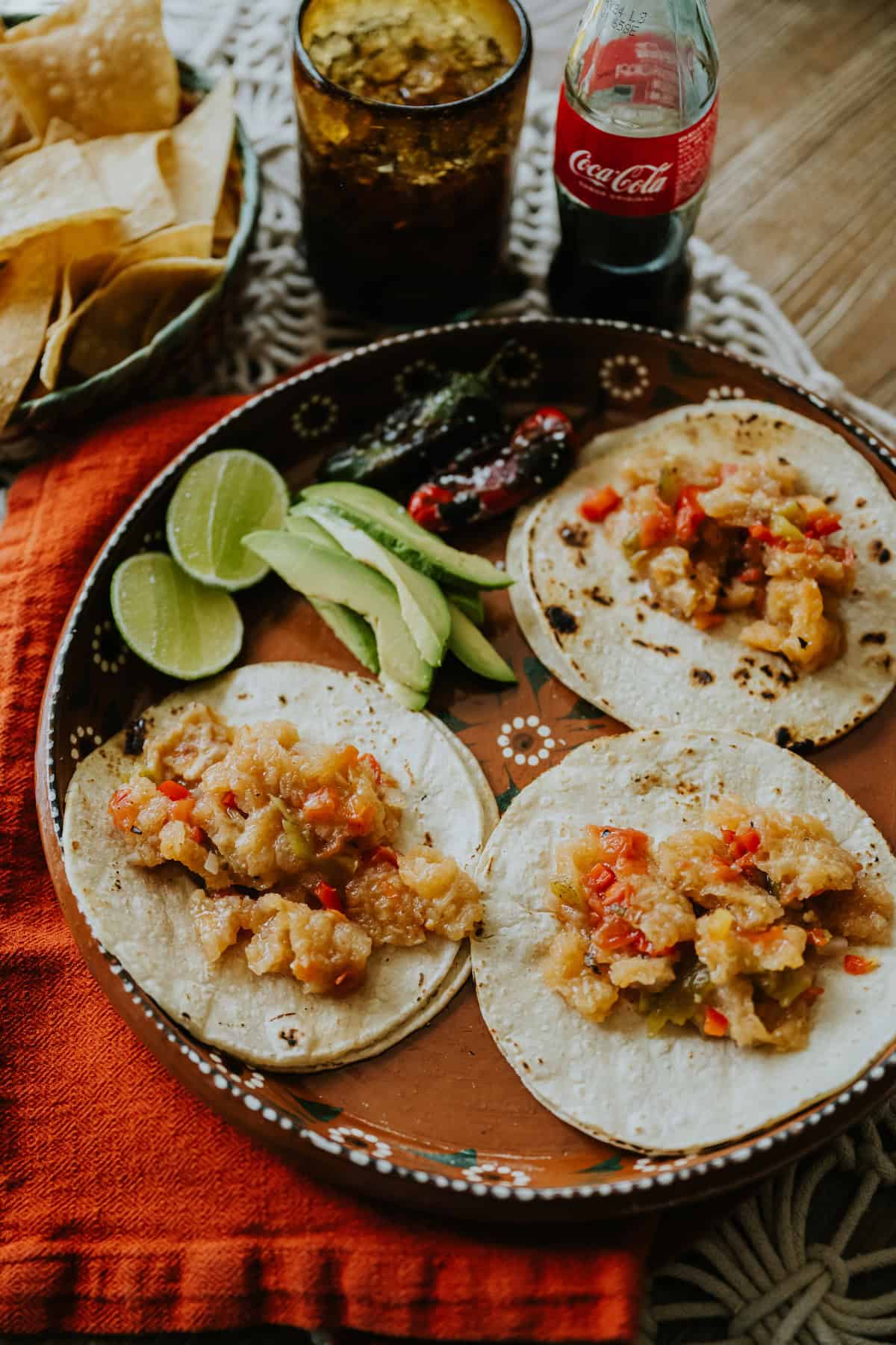 3 tacos de chicharron on a terracotta serving platter with sliced avocado, roasted jalapeños, and halved limes on a table next to a bottle of Mexican coke and a bowl of tortilla chips.