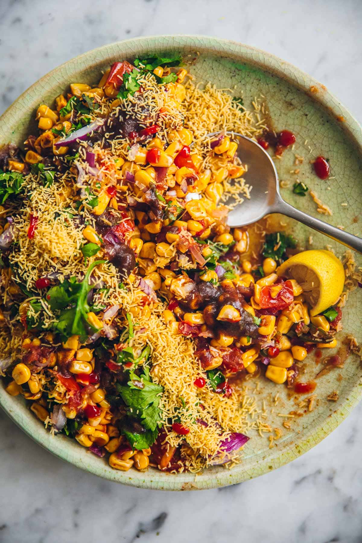 Corn Chaat served in a green plate topped with sev, onion, coriander, sweet date chutney and lemon wedge.