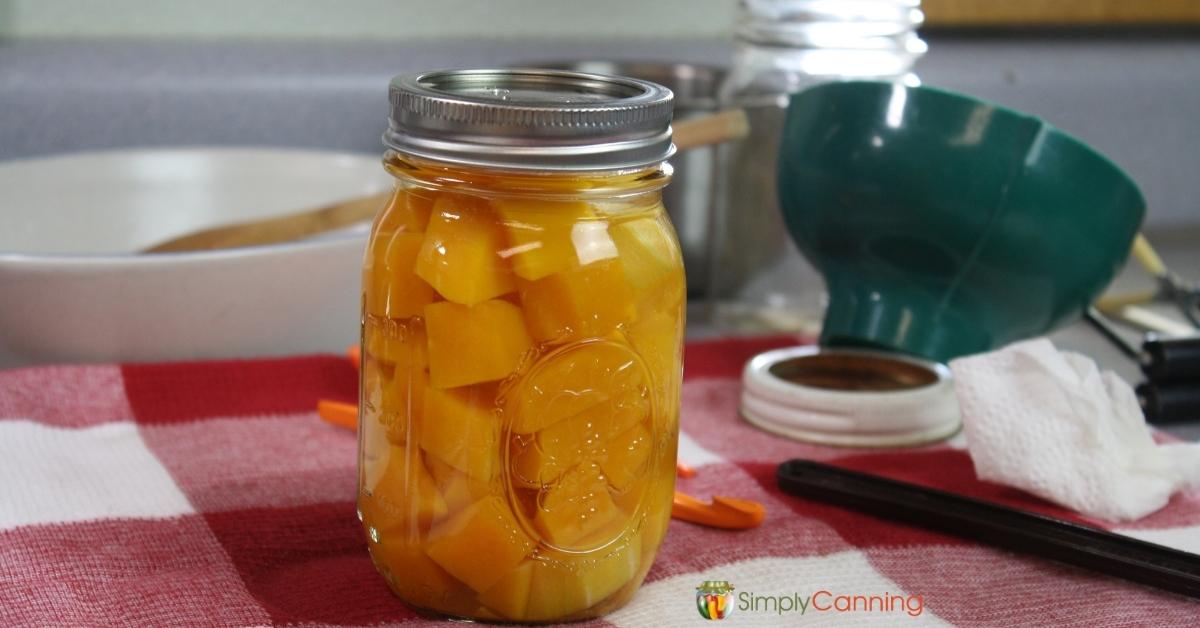 Pint jar of home canned butternut squash, sitting on a red checked towel with various canning tools in the background.