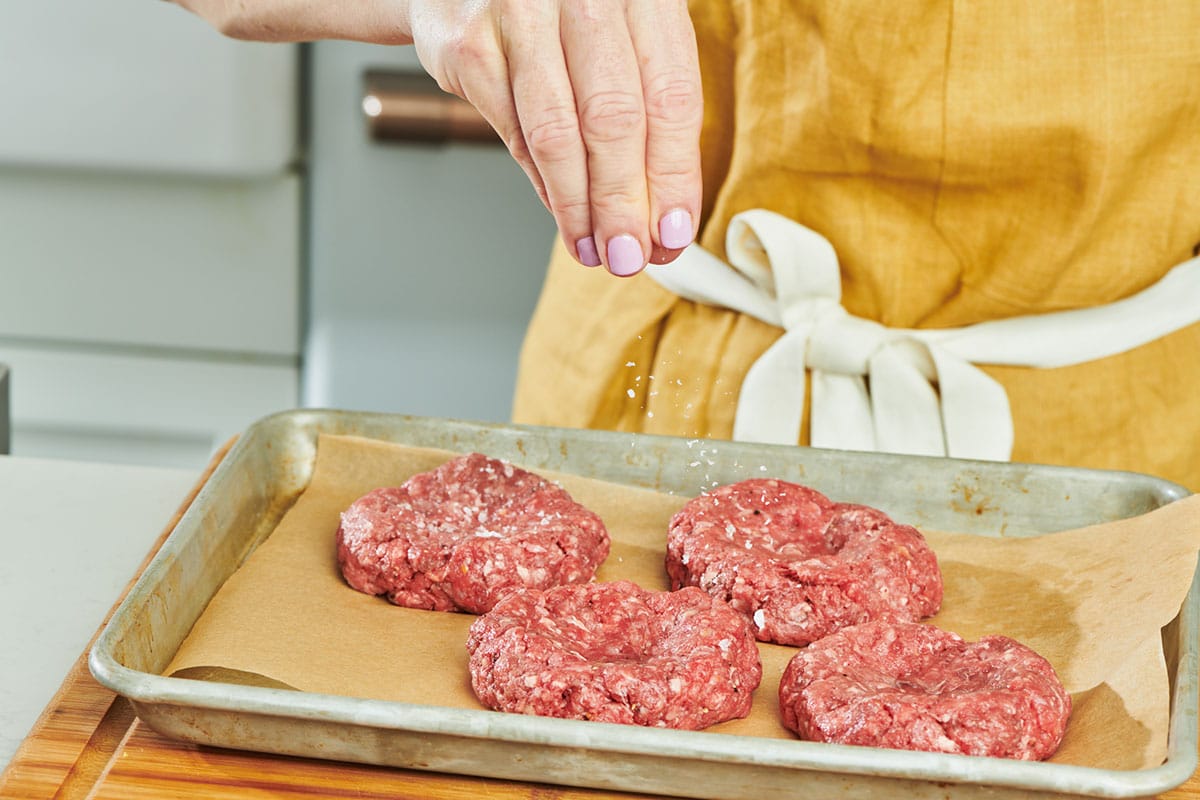 Woman sprinkling salt on raw hamburger patties.