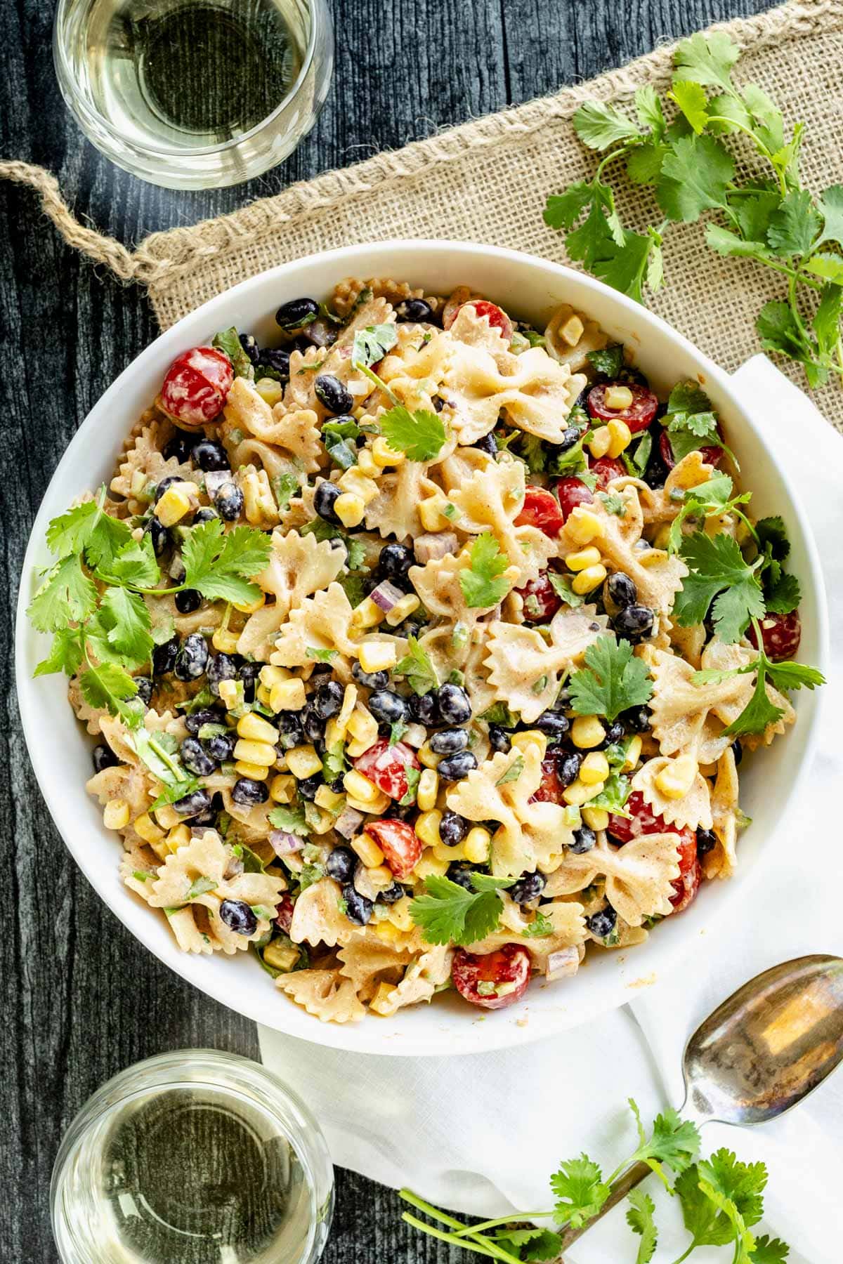 the bowl of Mexican pasta salad on a black table with a rustic mat underneath it with two glasses of white wine