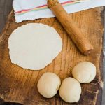 wooden cutting board with balls of dough and a rolled out tortilla next to a matching wooden palote.