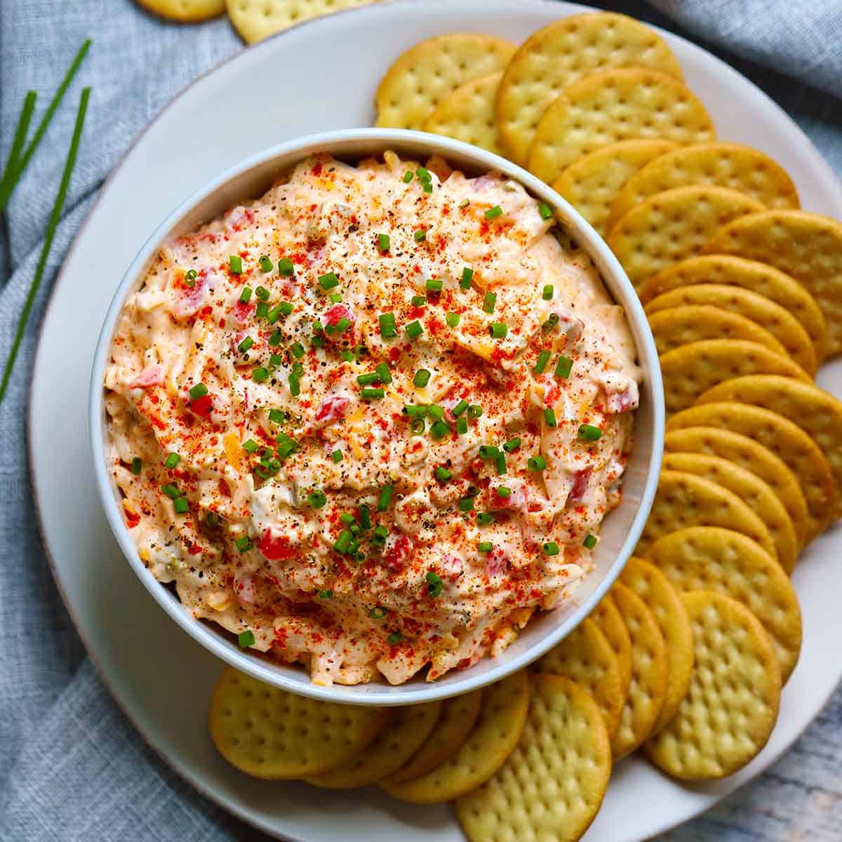 Square photo of overhead view of a bowl of pimento cheese surrounded by crackers on a plate.