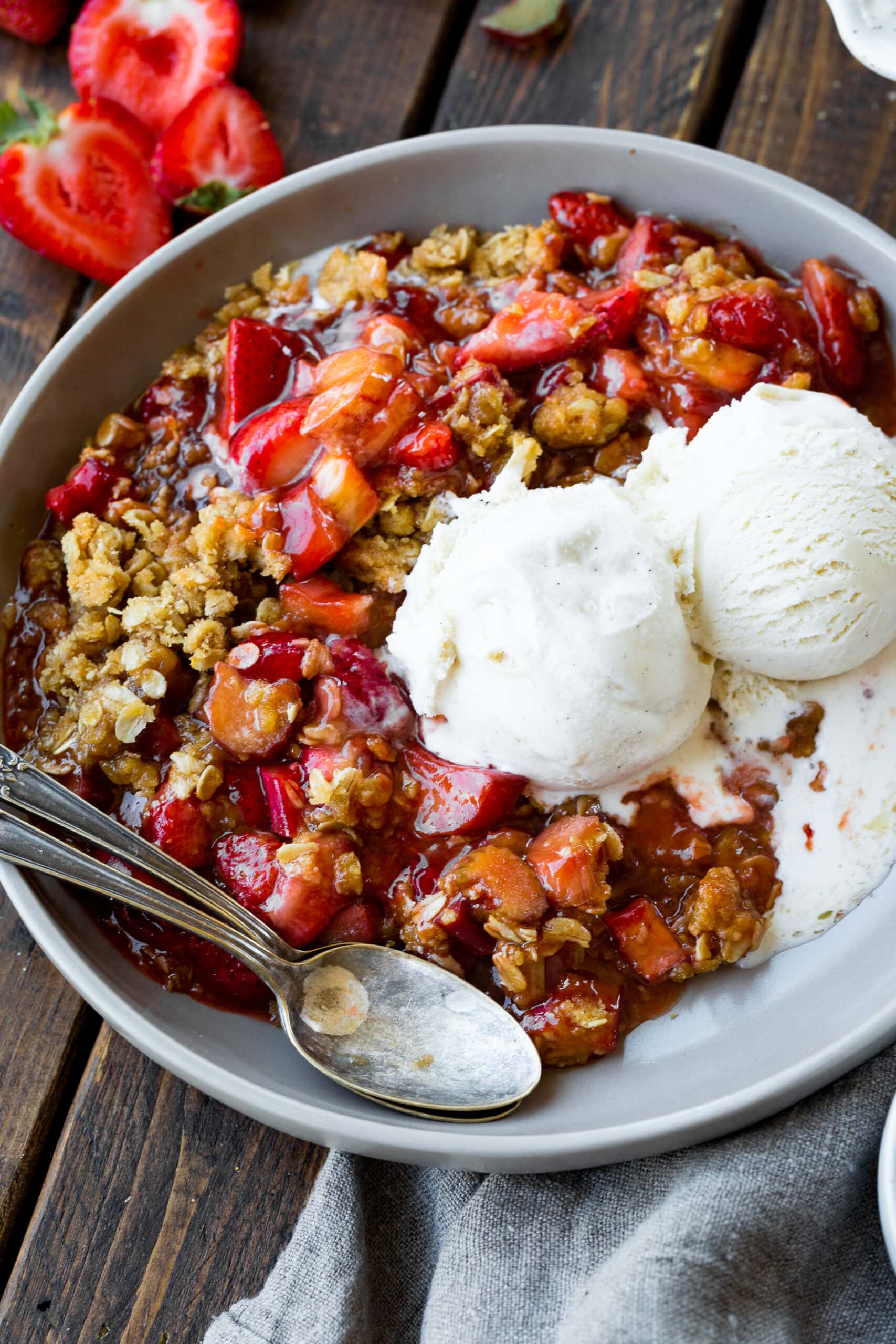 a photo of a serving of strawberry rhubarb crisp with a crunchy oat topping topped with two scoops of vanilla ice cream.