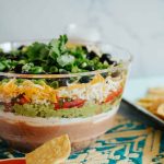 7 layer dip in a bowl on a Mexican textile with chips scattered.