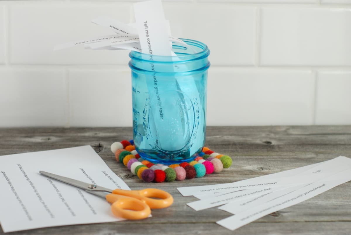 A blue jar of conversation starters sits on a wood table alongside a pair of scissors and slips of paper.