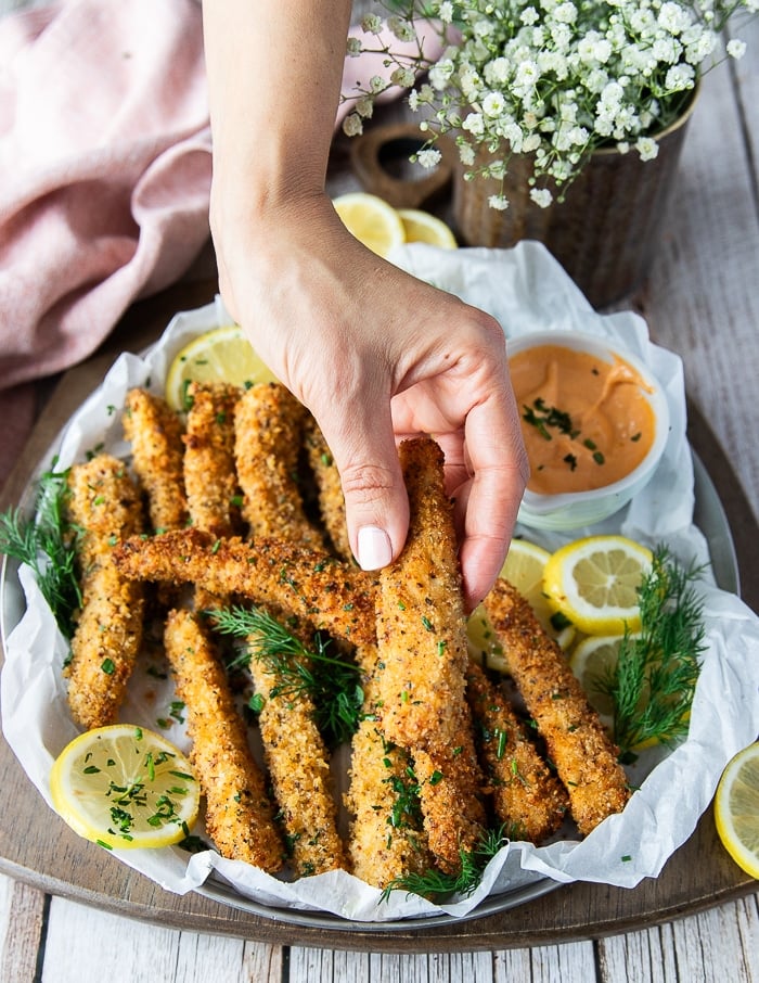 A hand holding one crunchy golden fish finger over a plate of fish fingers and some lemon slices