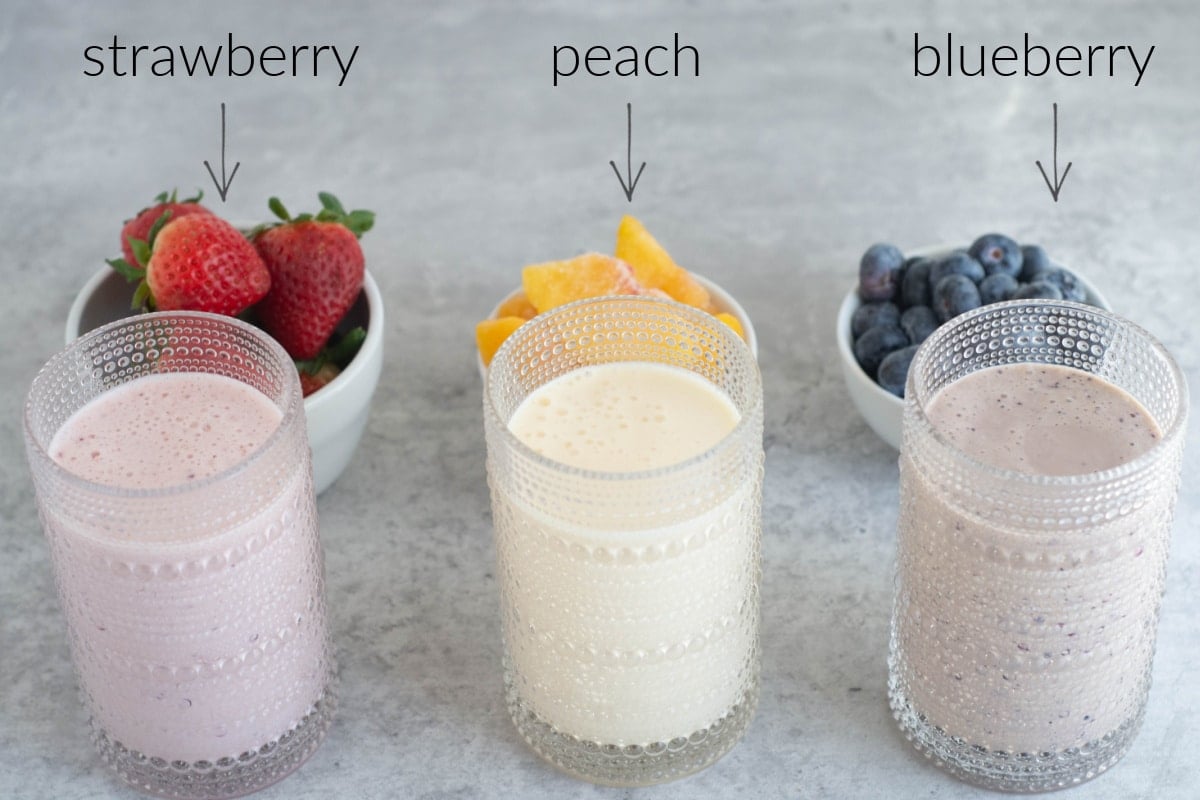 Glasses of drinkable yogurt sit in front of the fresh fruit that's in them: strawberries, peaches, and blueberries.