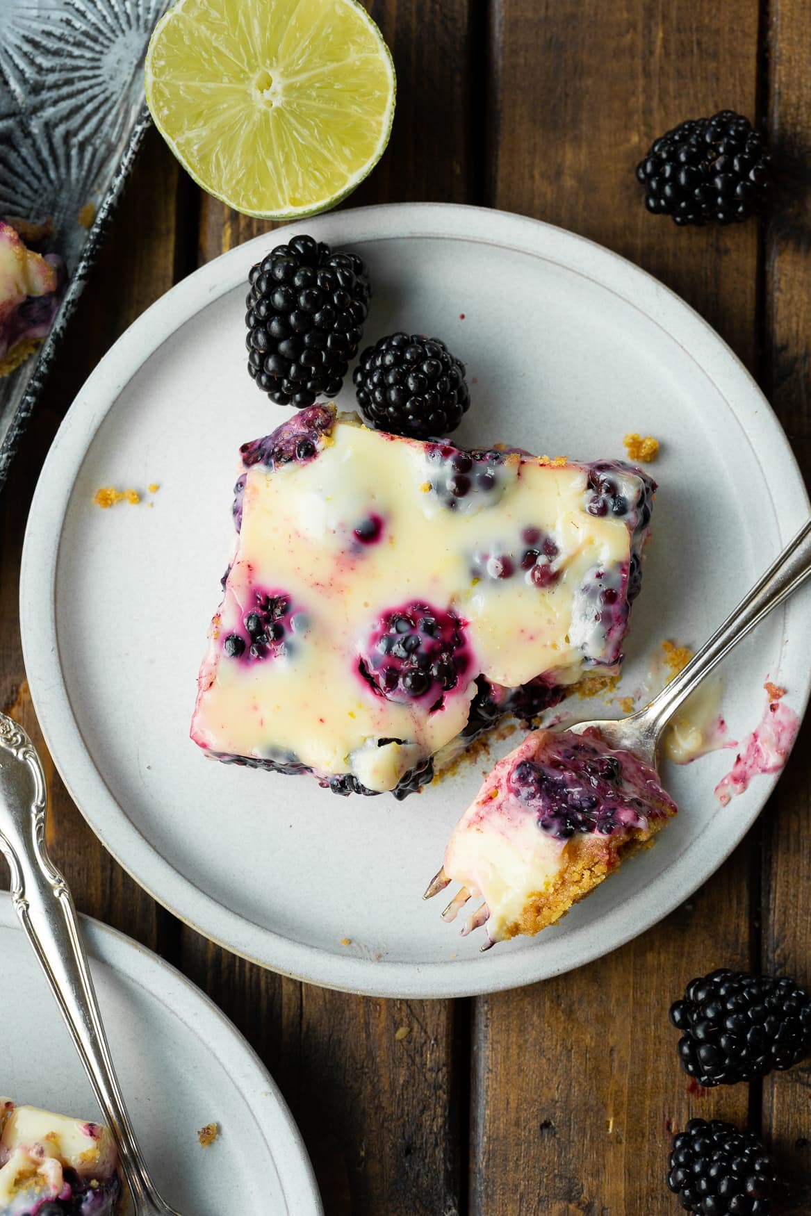 a photo of a blackberry lime bar sitting on a small plate with a corner scooped off by a fork sitting on the plate next to it.