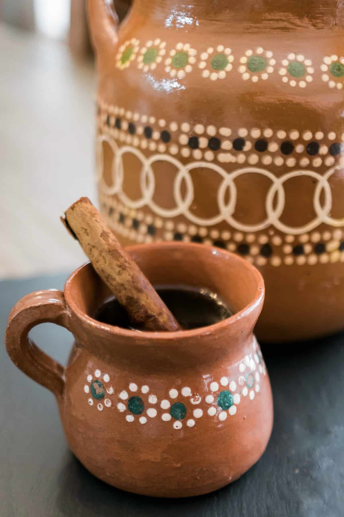 hand painted terracotta mug and olla filled with cinnamon tea, with the mug in front garnished with a canela cinnamon stir stick.