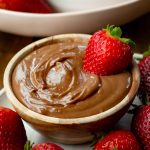 a photo of a small bowl filled with creamy chocolate yogurt dip with a whole strawberry dipped in it and strawberries scattered all around it