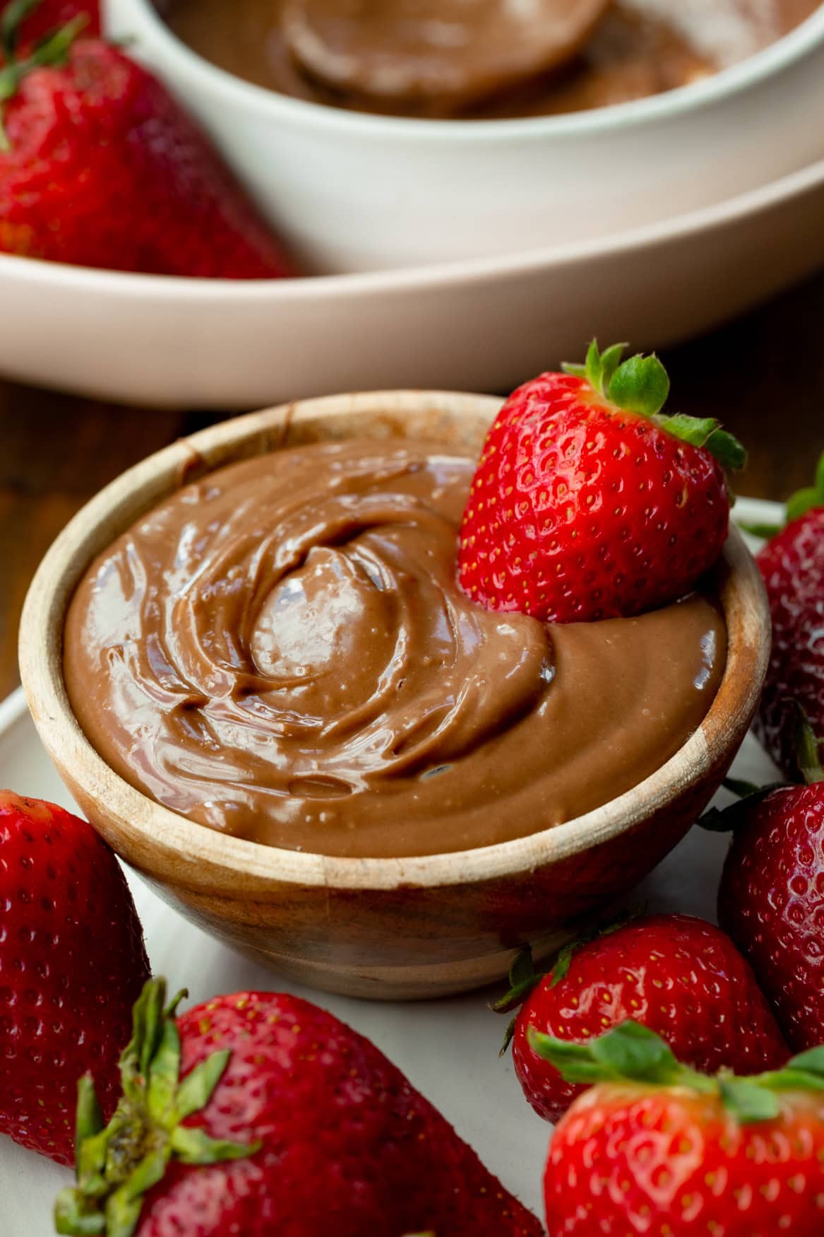 a photo of a small bowl filled with creamy chocolate yogurt dip with a whole strawberry dipped in it and strawberries scattered all around it