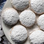 Square photo of overhead view of kourabiedes cookies on a white plate, coated in lots of powdered sugar.