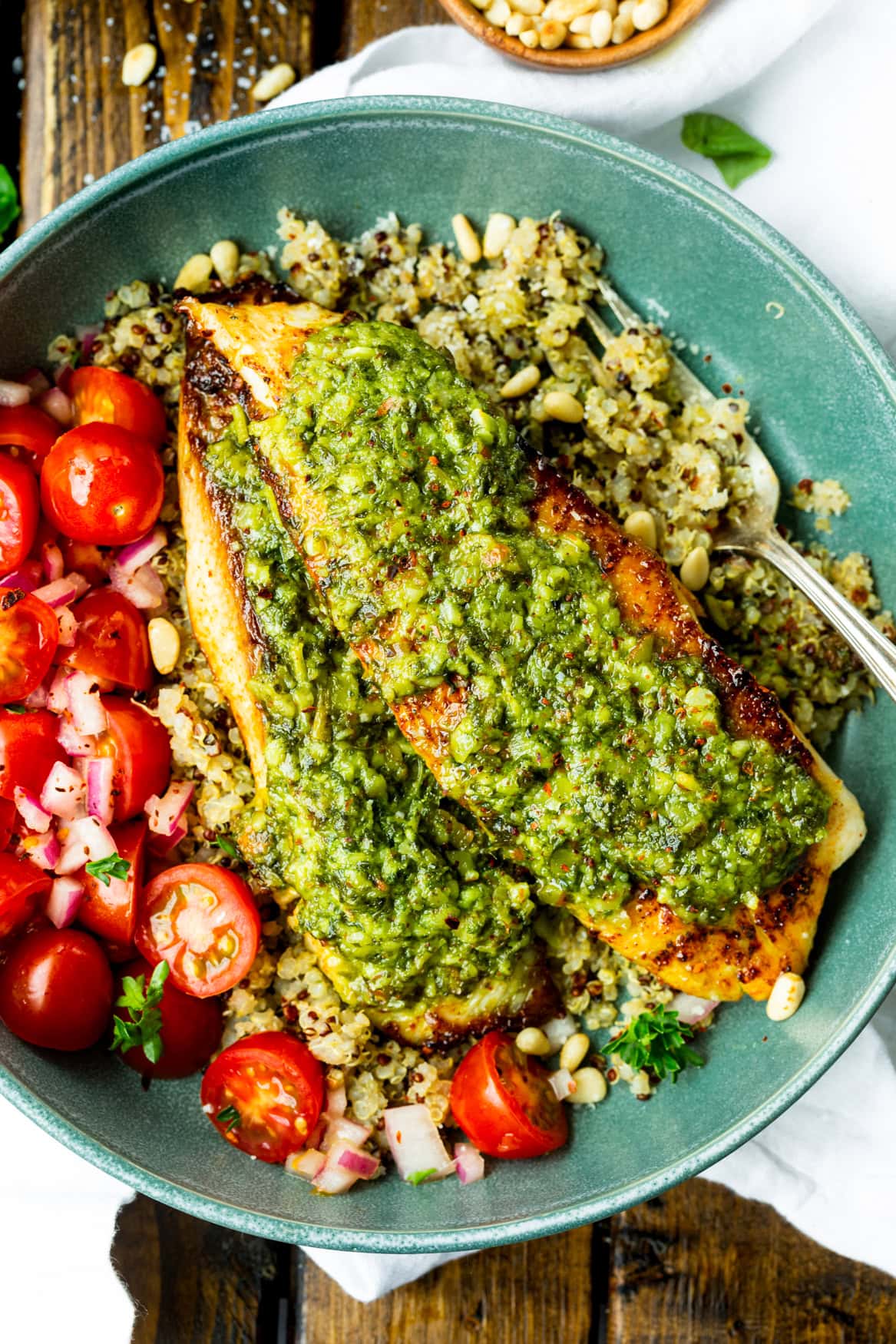 a photo of two golden tilapia filets topped with bright pesto sauce on a bed of quinoa with grape tomatoes on the side