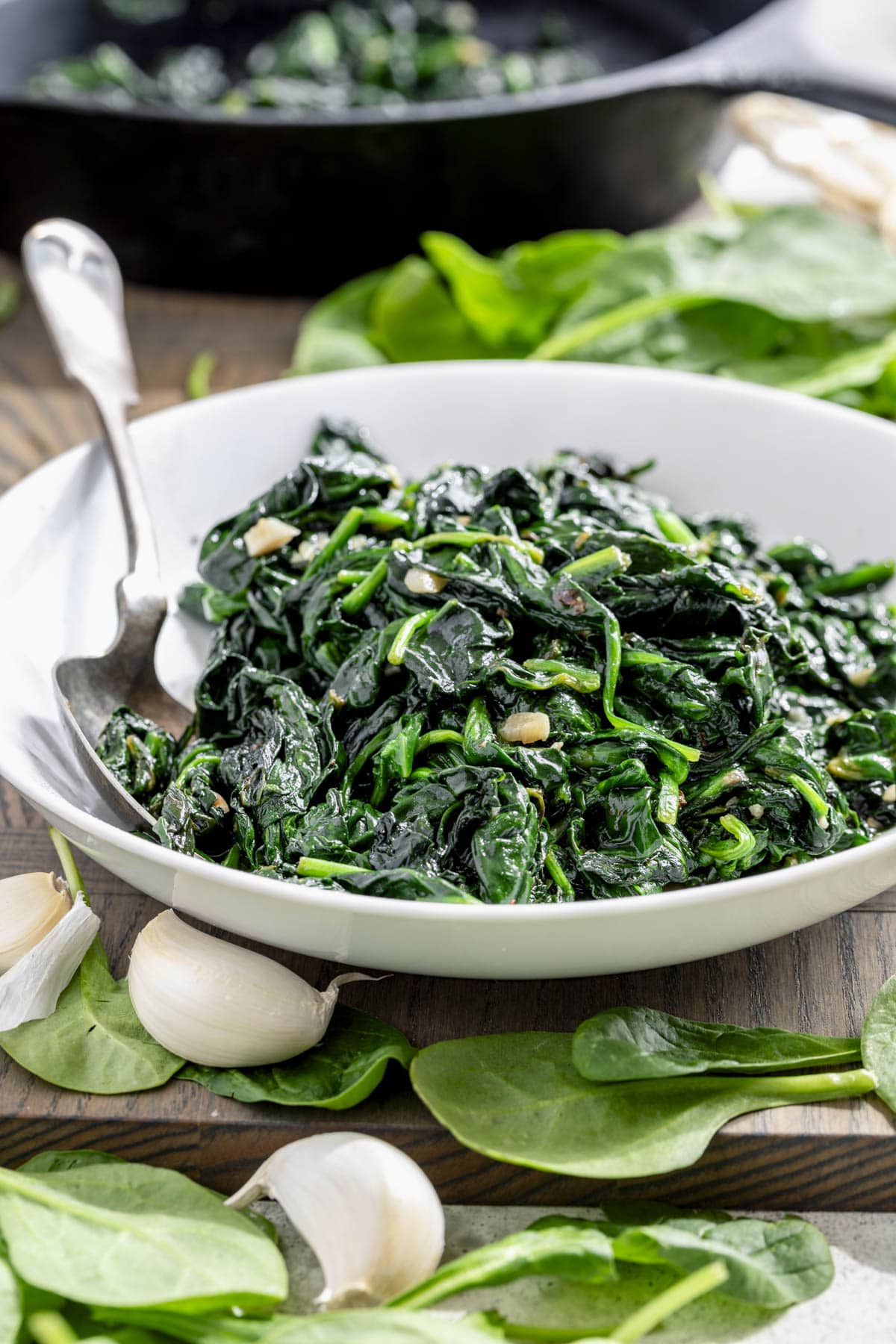 sauteed spinach in a white bowl with garlic cloves and fresh spinach leaves on the cutting board