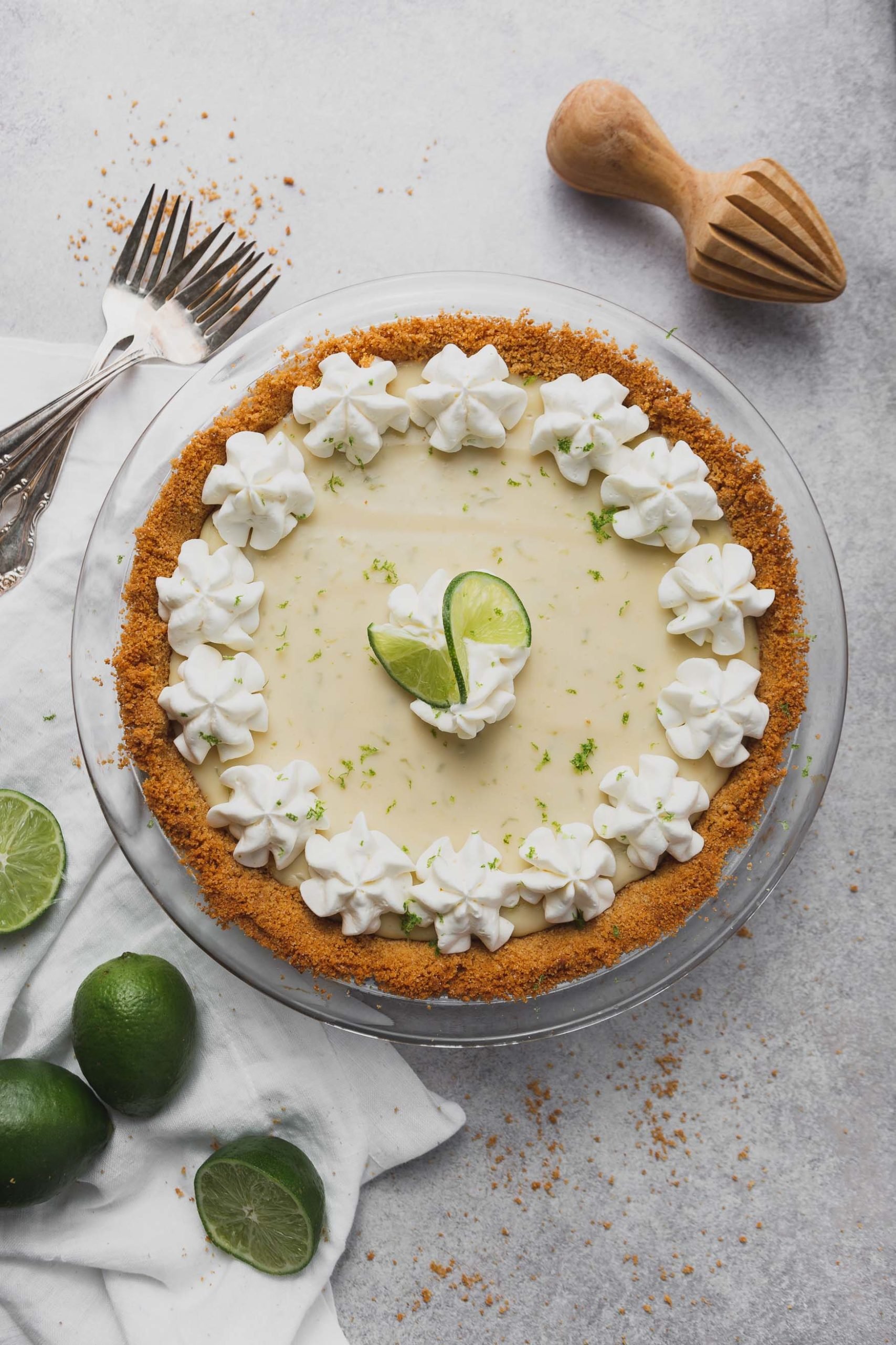 A key lime pie in a graham cracker crust. Dollops of whipped cream are around the edge of the pie and two slices of lime are in the middle. Limes and forks are beside the pie.