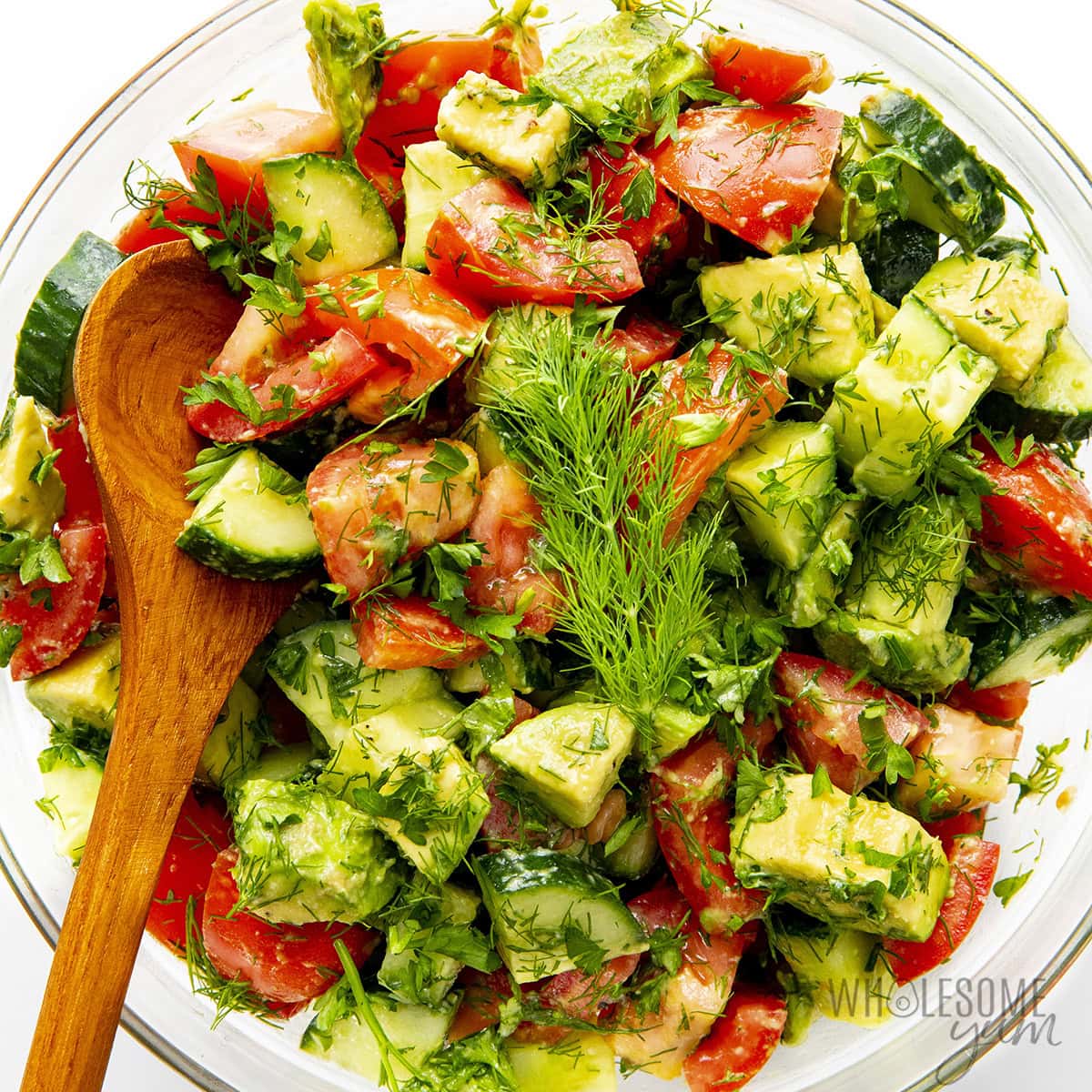 Cucumber tomato avocado salad in a serving bowl with a wooden spoon.